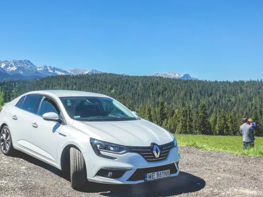 Biały Renault Megane Sedan zaparkowany na szutrowej drodze z widokiem na góry i las. Na trawie stoi mężczyzna z dzieckiem.