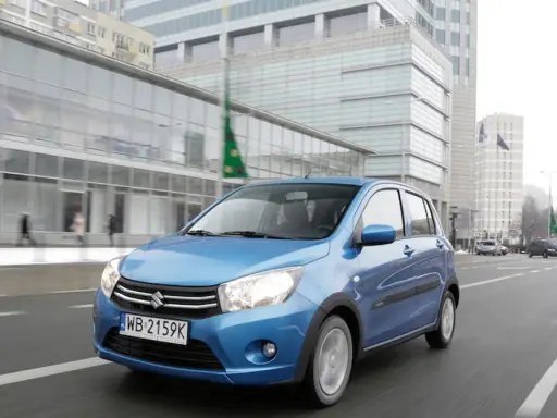 Niebieski Suzuki Celerio jedzie ulicą w mieście, mijając nowoczesne budynki i innych uczestników ruchu.