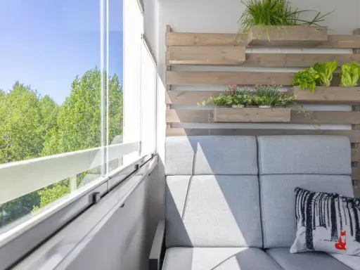 Przytulny balkon w bloku z szarą kanapą, poduszką z liskiem i zielenią na drewnianej ściance.