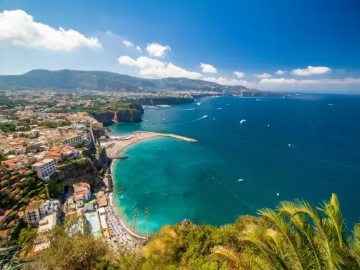 Vista panoramica della costa con il **scoglio del cavaliere** che emerge dal mare turchese, spiagge affollate e case aggrappate alla scogliera.