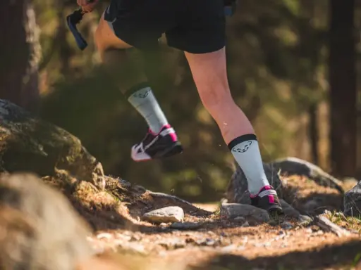 Beine einer Person beim **trail running** auf einem steinigen Waldweg. Leichte Sportschuhe und helle Socken mit Logo.