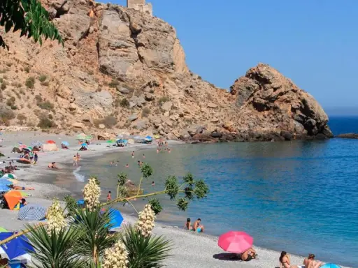 Playa de arena gris con gente disfrutando del sol y el mar, rodeada de acantilados. Un paraíso entre los pueblos de Granada con playa.