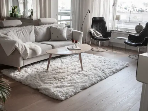 En fluffig, vit matta ligger framför en ljusgrå soffa och skapar en mysig känsla i vardagsrummet.