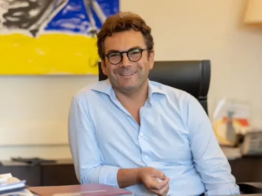 A man with glasses and a light blue shirt smiles while sitting at a desk with abstract art in the background.