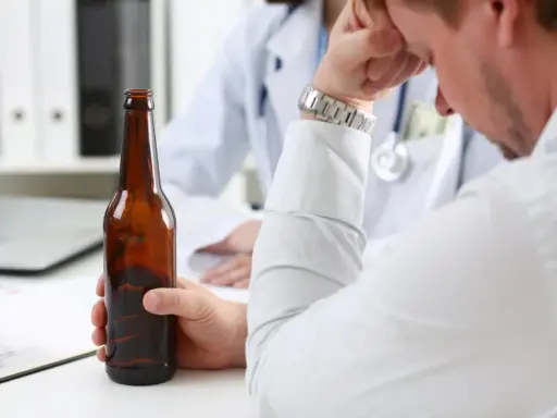 Mężczyzna trzyma butelkę piwa, siedząc przed lekarzem. Rozmowa sugeruje skierowanie na detoks alkoholowy.