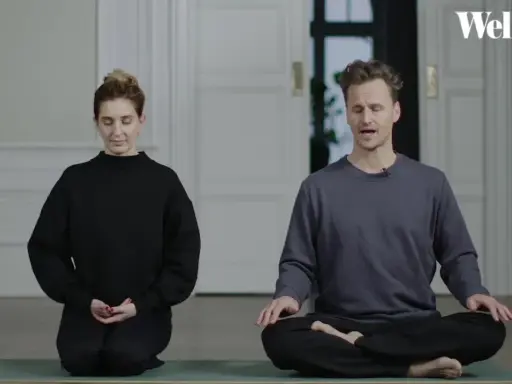 Två personer sitter på en yogamatta och mediterar. En kvinna i svart tröja och en man i grå tröja. Ta ett ögonblick att stanna upp.