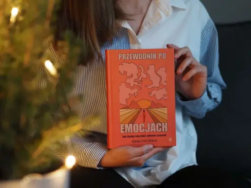 Osoba trzyma pomarańczową książkę "Przewodnik po emocjach". Okładka przedstawia abstrakcyjne kształty i zachód słońca.