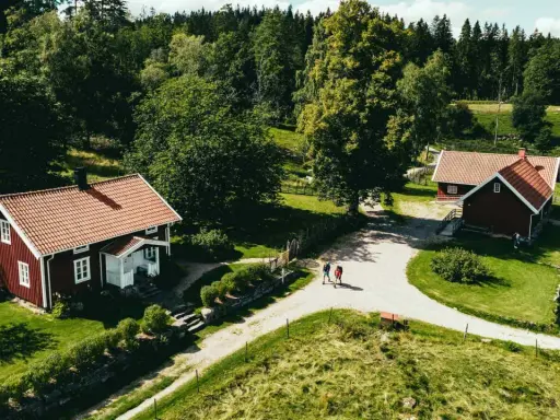 Två röda hus med tegeltak vid Rya Åsar, Borås. En grusväg leder förbi husen genom ett grönskande landskap med skog och ängar.