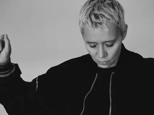Jennifer Cardini, con i suoi capelli biondi corti e un'espressione pensierosa, alza un braccio con un gesto delicato. Indossa una giacca scura con zip.