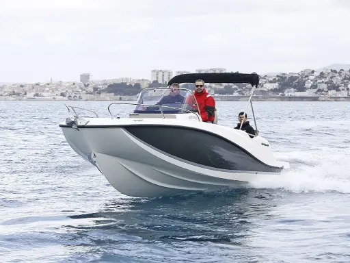 Bateau blanc et gris naviguant sur l'eau, avec trois personnes à bord. Le tableau de bord indique la puissance du moteur du bateau. Ville en arrière-plan.