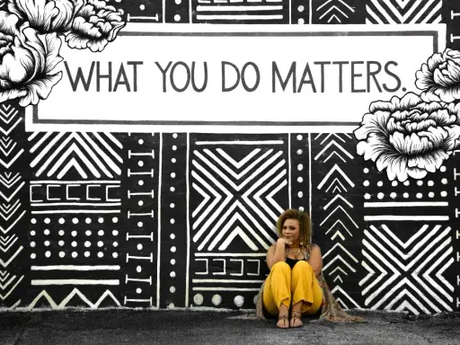 Kobieta siedzi przed muralem z napisem "WHAT YOU DO MATTERS". To inspirujący dialog motywujący.