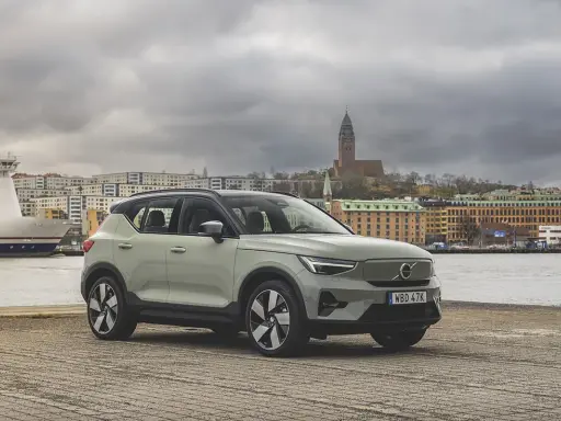 A light green Volvo XC40 parked by a waterfront with a ferry and city buildings in the background.