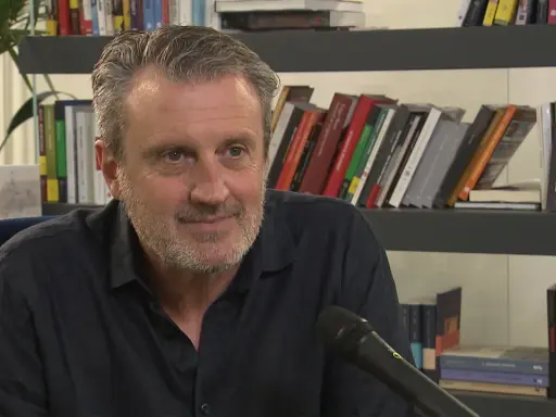 Uomo con barba grigia sorride, microfono davanti. Alle sue spalle scaffali pieni di libri, tra cui "Le ossa parlano trama".
