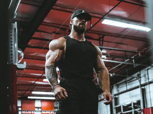 Ein Mann mit beeindruckendem **mens physique** steht im Fitnessstudio. Seine Muskeln sind stark definiert, er trägt eine schwarze Kappe und ein Tanktop.