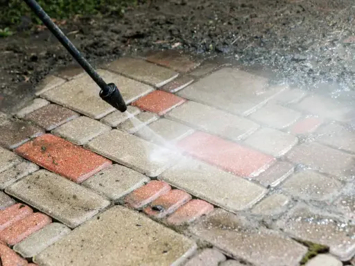 Czyszczenie kostki brukowej myjką ciśnieniową. Woda usuwa brud z czerwonych i szarych płyt.
