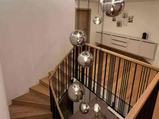 Nowoczesna klatka schodowa w domu z drewnianymi stopniami i czarną balustradą, ozdobiona wiszącymi, lustrzanymi kulami.