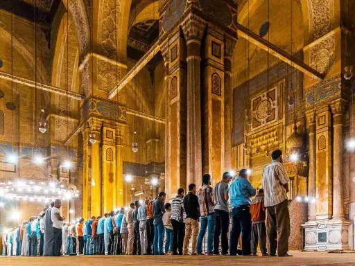 Männer beten Tarawih in einer Moschee. Sie stehen in Reihen vor kunstvoll verzierten Säulen und Bögen, beleuchtet von zahlreichen Lampen.