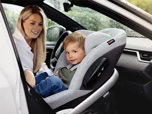 Uśmiechnięta mama patrzy na dziecko w foteliku z przodu. Widoczna jest poduszka powietrzna pasażera.