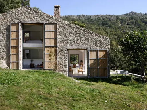 Una casa in pietra con grandi porte in legno, perfetta per chi ama vivere in campagna. Si intravedono interni moderni e un paesaggio collinare rigoglioso.