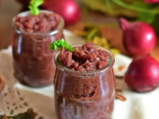 Dwa słoiczki z domowym, aromatycznym chutney z czerwonej cebuli, ozdobione listkiem zieleniny.