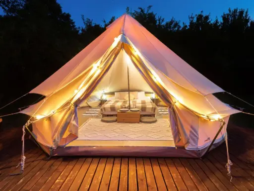 Mysig glamping i Sverige. Ett upplyst tält med säng och mysig inredning på en träaltan, omgivet av mörka träd.