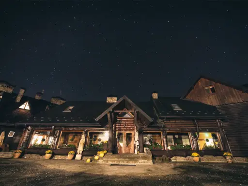 Nocne niebo nad bacówką Biały Jeleń. Drewniany budynek z oknami oświetlonymi ciepłym światłem, otoczony donicami z kwiatami.