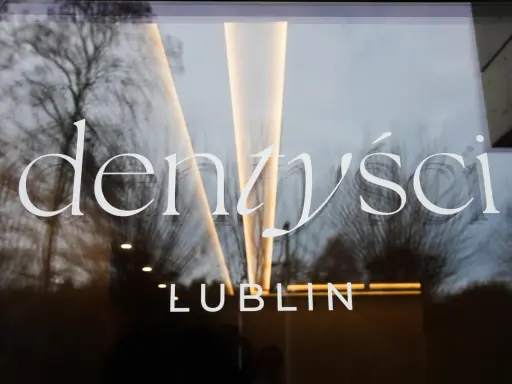 Napis "Dentyści Lublin" na szklanych drzwiach z odbiciem drzew i światła.