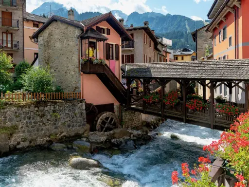 Un pittoresco ponte di legno attraversa un torrente impetuoso, fiancheggiato da case colorate e fiori rossi. Cosa vedere in questo incantevole borgo alpino.