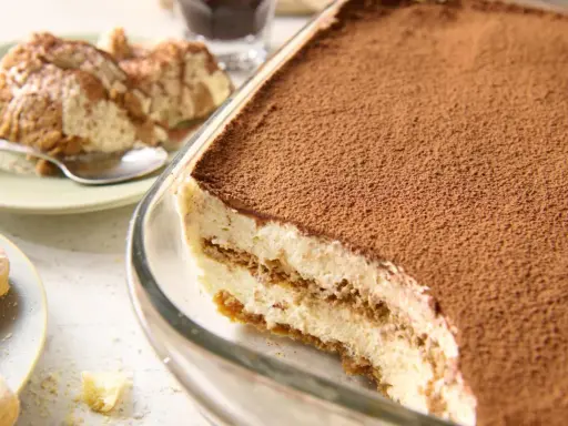Pyszne tiramisu bez jajek, obficie posypane kakao, w szklanej formie. Obok porcja na talerzyku.