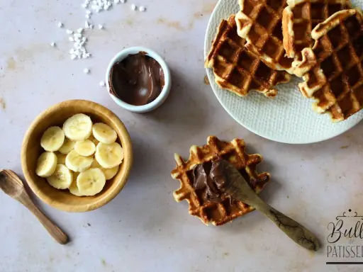Une gaufre bubble est garnie de pâte à tartiner au chocolat. À côté, des rondelles de banane et une cuillère en bois.