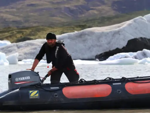Un homme tente de démarrer un moteur 2 temps sur un Zodiac, mais il cale. Derrière lui, des icebergs flottent dans un lac glaciaire.