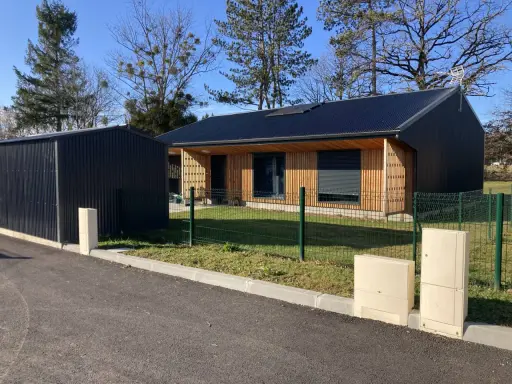 Maison passive ossature bois avec panneaux solaires sur le toit, bardage bois et métal, devant un ciel bleu et des arbres.