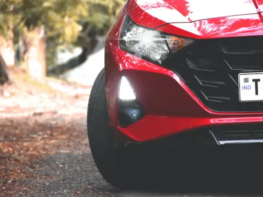 Czerwony Hyundai i20 z zapalonymi światłami dziennymi i reflektorami. Widać fragment maski, grill i tablicę rejestracyjną.