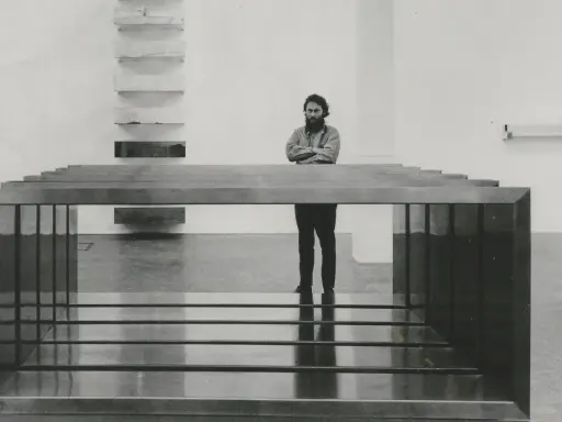 A man stands with arms crossed, contemplating a minimalist installation of Donald Judd art, featuring stacked shelves and a long, rectangular sculpture.