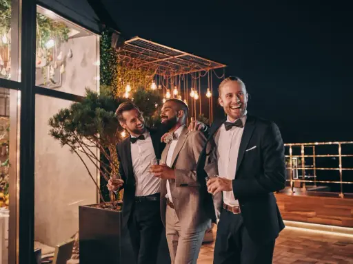 Trzech eleganckich panów, w tym drużba na weselu, śmieje się i trzyma drinki na tarasie nocą, wśród ozdobnych świateł.