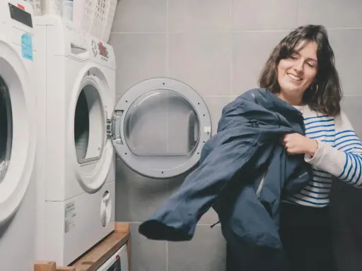 Kobieta z uśmiechem trzyma granatową kurtkę, przygotowując się do prania Gore-Tex. Obok stoi otwarta suszarka.