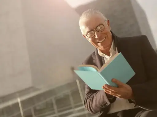 Ein älterer Mann mit Brille lächelt und hält ein hellblaues Buch. Er scheint Freude am Auswendiglernen von Gedichten zu haben.