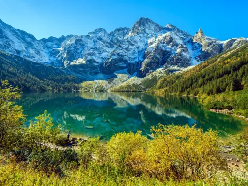 Widok na Morskie Oko z wysokości. Turkusowa tafla jeziora odbija ośnieżone szczyty Tatr. Jesienne drzewa otaczają brzeg.