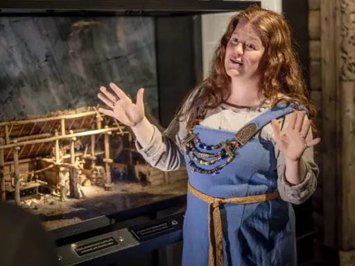 En kvinna i vikingakläder berättar om en modell av en vikingagård på ett vikingamuseum.