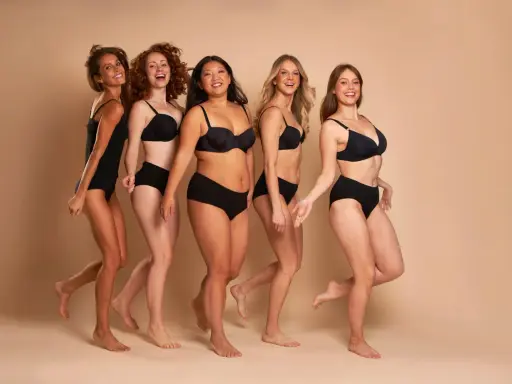 Cinque donne diverse, con taglia nona di seno e forme varie, camminano sorridenti in lingerie nera.