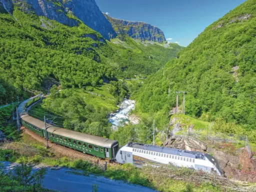 Grüner Zug fährt durch ein bergiges Tal mit Fluss. Die Fahrt von Bergen nach Oslo ist ein unvergessliches Erlebnis.