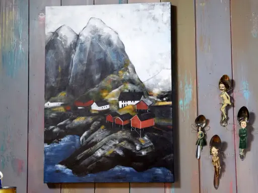 Målade landskap med röda hus vid en bergig kust. Konstverket hänger på en vägg av slitna träplankor.