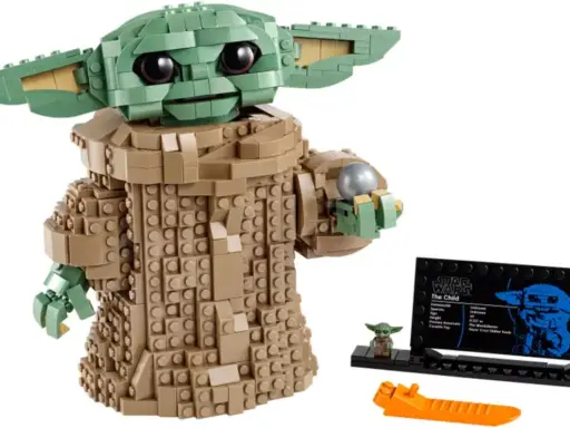 Figurka Grogu z klocków LEGO, znana z gier LEGO Star Wars, trzyma srebrną kulę. Obok stoi mała figurka i tabliczka informacyjna.