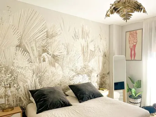Dormitorio con papel pintado de palmeras, ideal para aprender como poner papel pintado. Cama con cojines oscuros, espejo y planta.