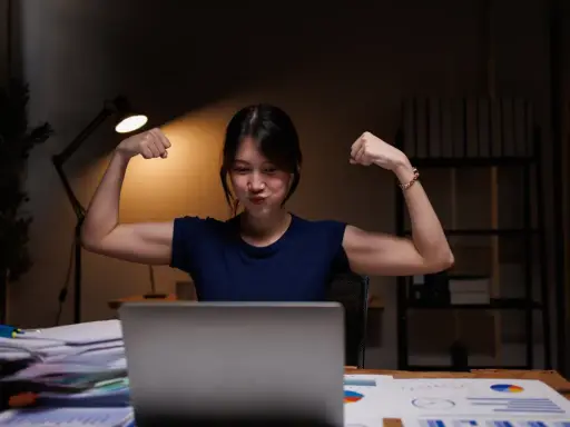 Młoda kobieta w godzinach nocnych 2026, siedząc przy biurku, pokazuje bicepsy.