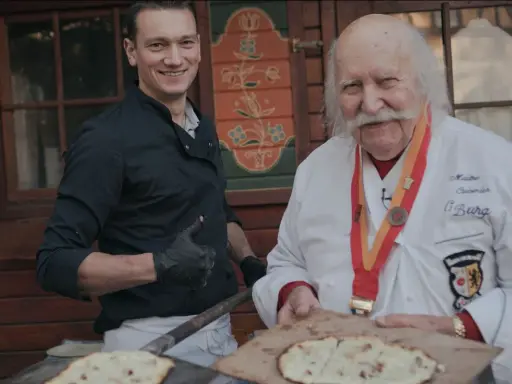 Deux chefs, l'un jeune et souriant, l'autre âgé avec une médaille, présentent des tartes flambées, évoquant l'origine du flamenkuch.