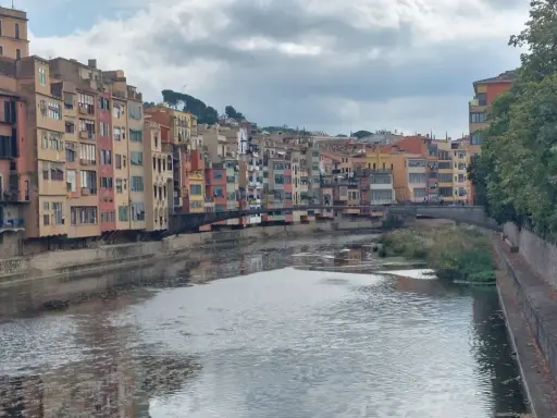 Bunte Häuserfronten säumen den Fluss Onyar in der malerischen **Girona Altstadt**. Eine Brücke verbindet die Ufer, während sich die Wolken am Himmel spiegeln.