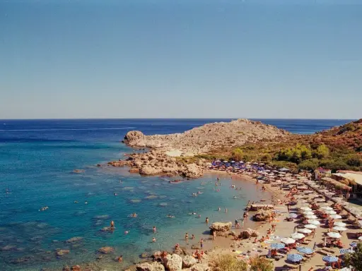 Rodos: turkusowe morze Egejskie, kamienista plaża z leżakami i parasolami, ludzie kąpiący się w wodzie.