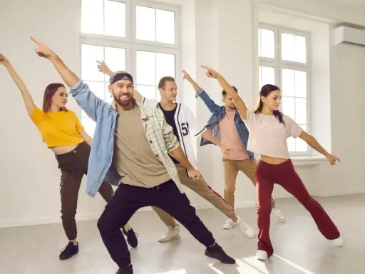 Grupa ludzi ćwiczy energiczne samba kroki w jasnej sali tanecznej, z rękami uniesionymi w rytm muzyki.