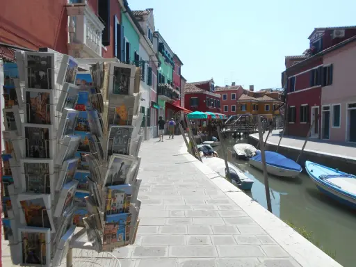 Cartoline di Murano, Burano e Torcello in vendita lungo un canale con case colorate e barche ormeggiate.